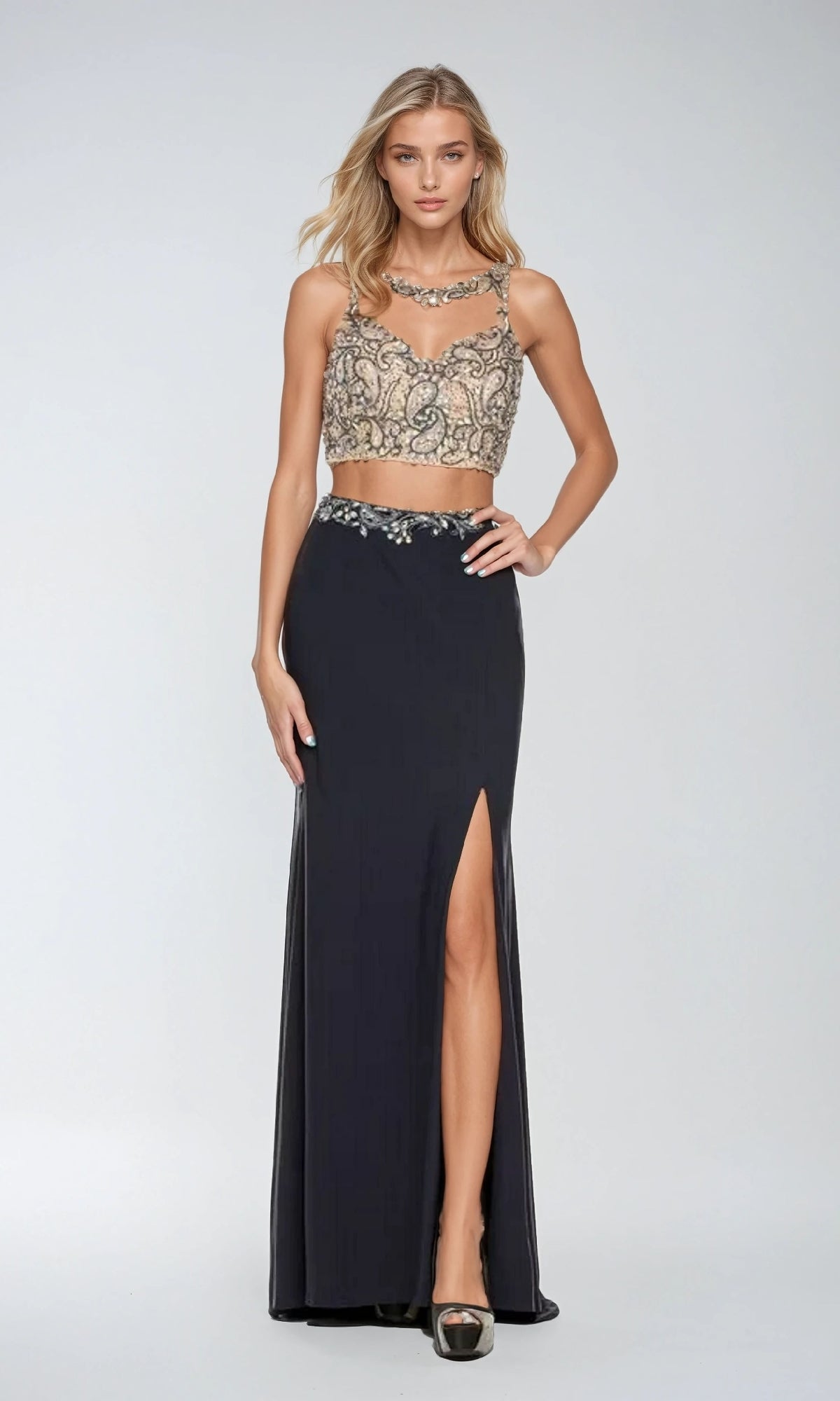 A woman wearing the Juliet 595 Long Prom Dress, featuring a beaded sleeveless crop top and a black skirt with a side leg slit, stands against a plain light background.